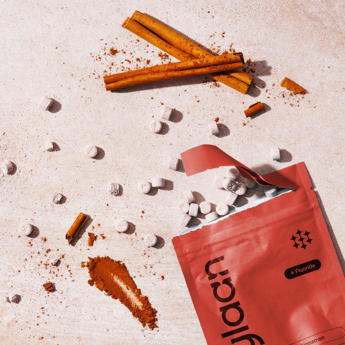 Top-down view of an orange Kaylaan Cinnamon toothpaste refill bag with loose tablets, cinnamon sticks, and ground powder scatter.