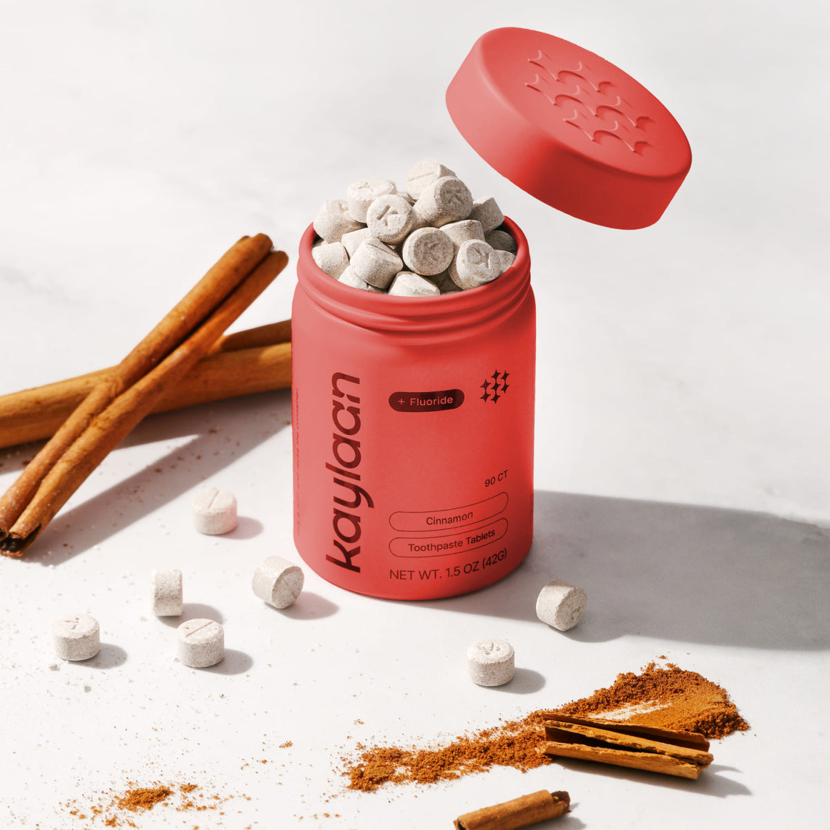 An open, vibrant red aluminum tin of Kaylaan Cinnamon Toothpaste Tablets with Fluoride, overflowing with small round white tablets stamped with a K. The matching red lid floats slightly above, surrounded by whole cinnamon sticks on a clean white surface.