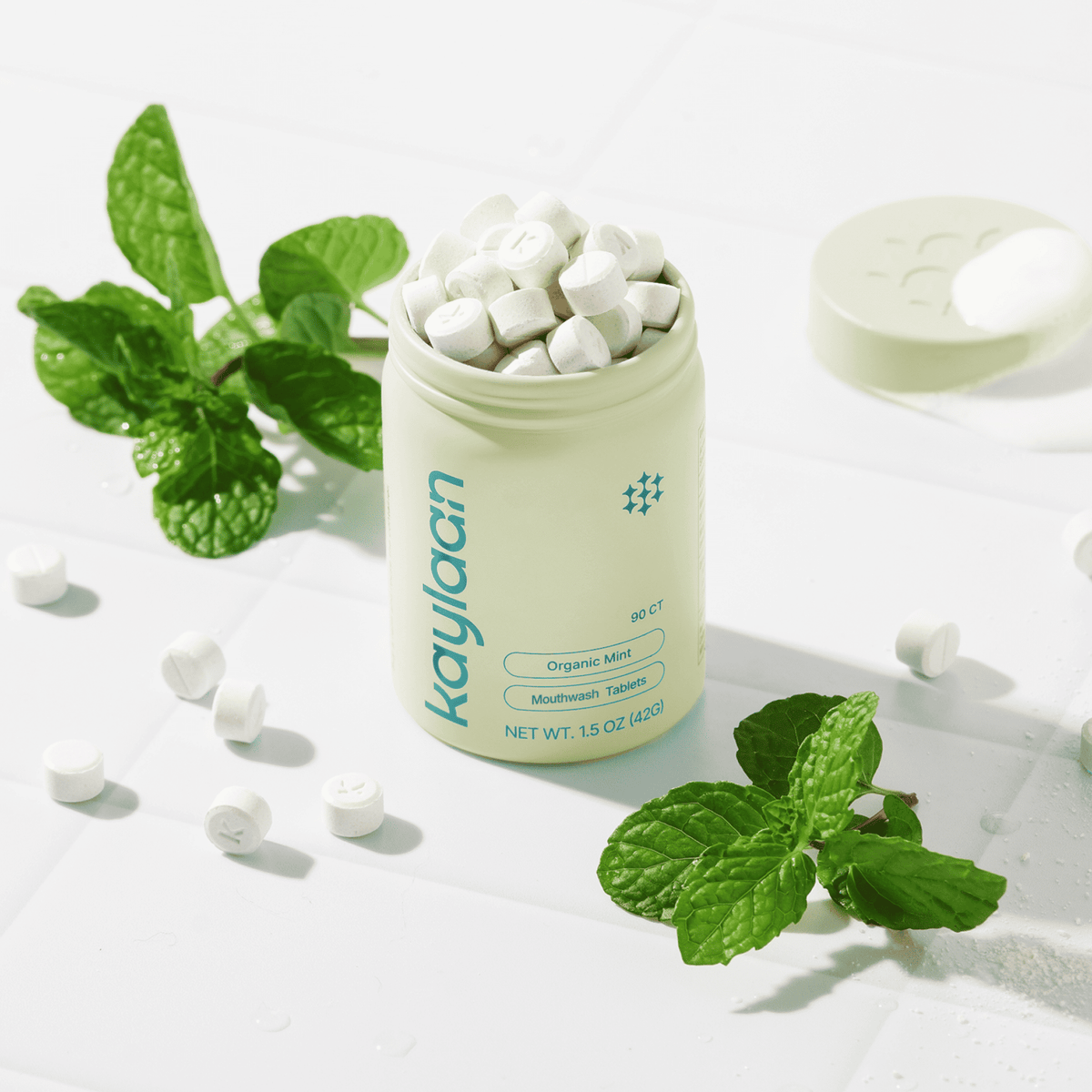 Overhead view of a light green tin overflowing with white Kaylaan mouthwash tabs, surrounded by loose tablets and fresh green mint leaves on a bright tile surface.