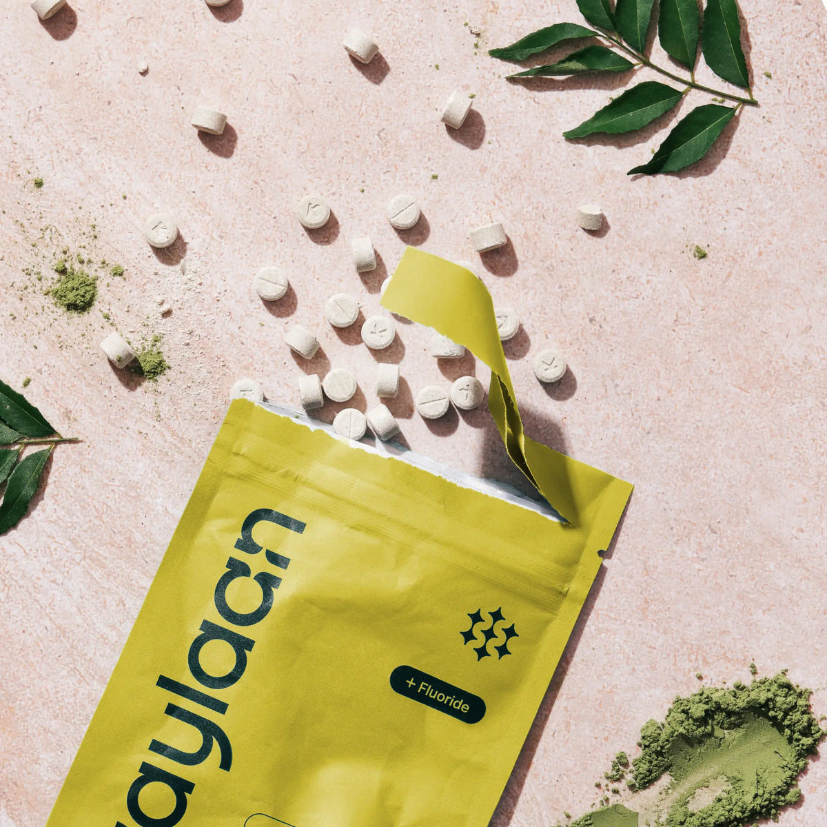 Overhead view of an open yellow Kaylaan Neem toothpaste refill pouch spilling tablets onto a textured pink surface with green leaves.