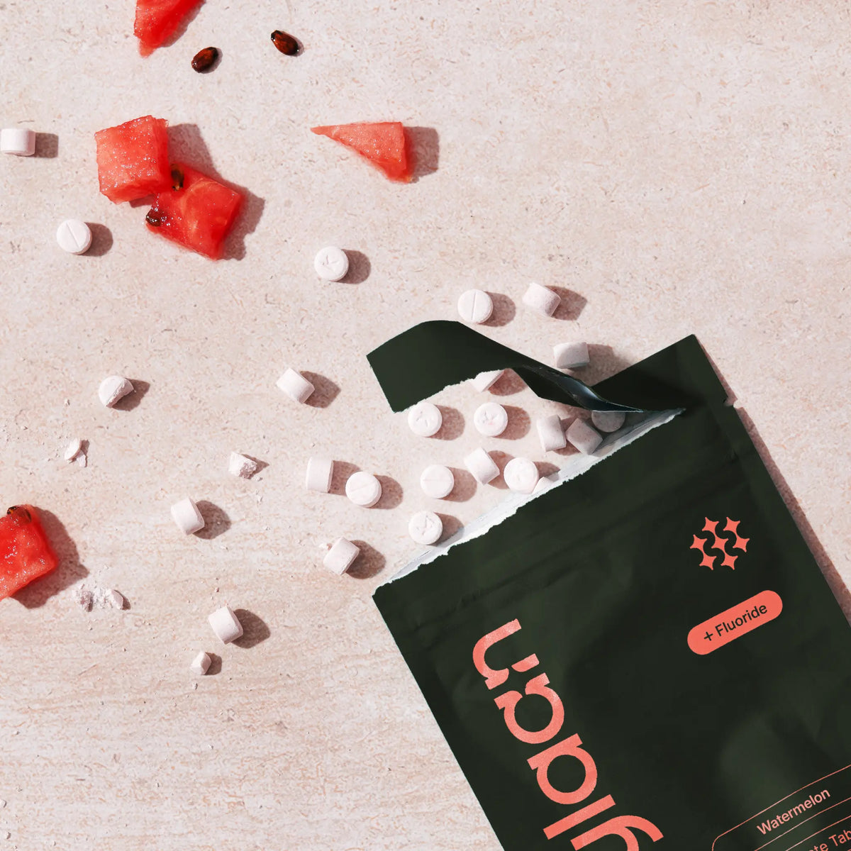 Overhead shot of a dark green Kaylaan Watermelon toothpaste tablet pouch spilling small white tabs alongside fresh watermelon pieces.